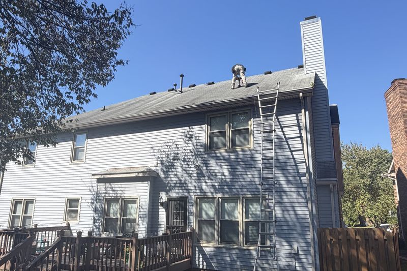An adjuster kneeling on the gray shingle roof of a two-story house with blue siding, with an extension ladder against the wall