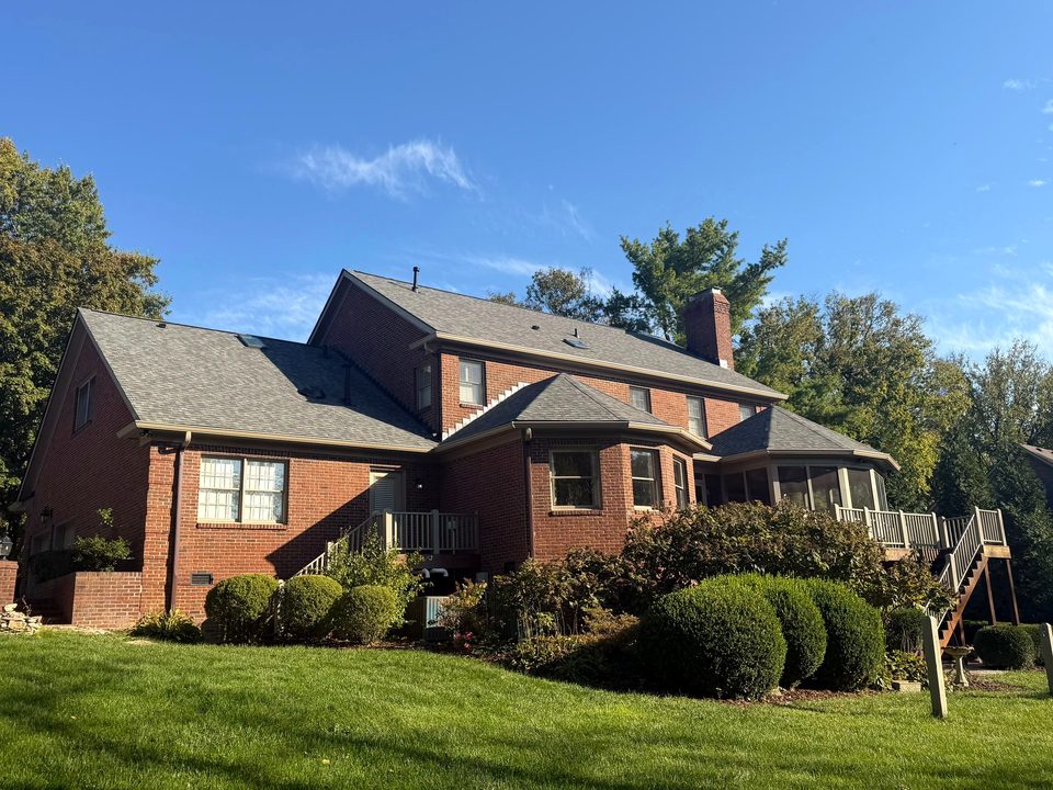 Rear of a two-story brick home with a new gray shingle roof, new two-tone gutters and flashing, a screened porch and deck, under a blue sky
